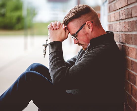 A man sitting alone, head bowed in deep thought or prayer against a brick wall, symbolizing the need for a reliable, easy-to-use, and spiritual platform to connect people with their faith.