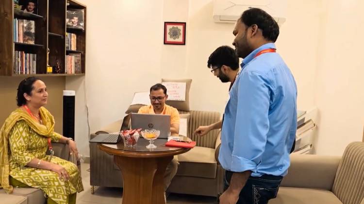 Four team members gathered in an office lounge, with one man working on a laptop at a round wooden table while the others strategize.